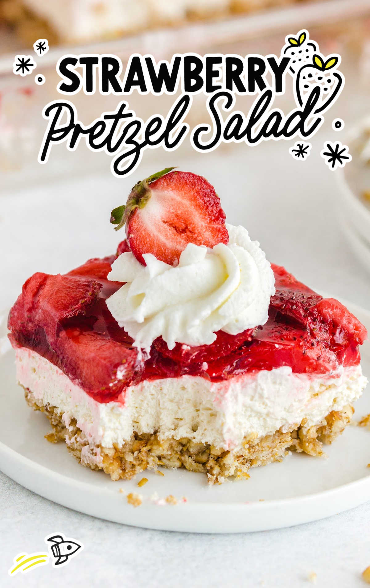 A slice of strawberry pretzel salad topped with whipped cream and a strawberry half sits on a white plate. This classic treat, featured in 28+ Classic Dessert Salads & Fluff Recipes Everyone Loves, boasts a pretzel crust and glazed strawberry topping.