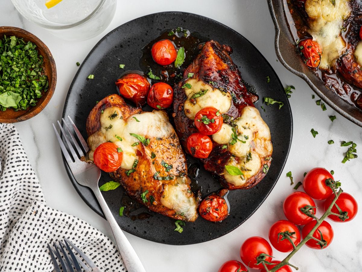 Balsamic Chicken with melted mozzarella and roasted cherry tomatoes served on a black plate with fresh basil.