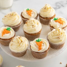 Twelve Carrot Cake Cupcakes with white frosting, some topped with small carrot decorations and others with chopped nuts, are arranged on a white cake stand—an irresistible treat inspired by a classic Carrot Cake Cupcakes Recipe.