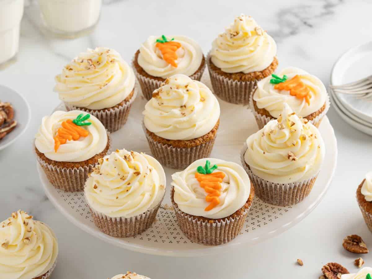 Twelve Carrot Cake Cupcakes with white frosting, some topped with small carrot decorations and others with chopped nuts, are arranged on a white cake stand—an irresistible treat inspired by a classic Carrot Cake Cupcakes Recipe.