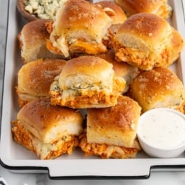 A tray of baked Buffalo Chicken Sliders filled with shredded buffalo chicken and melted cheese, topped with golden brown buns. A small dish of ranch dressing and a bowl of blue cheese crumbles are served on the side. Try this Buffalo Chicken Sliders Recipe!.