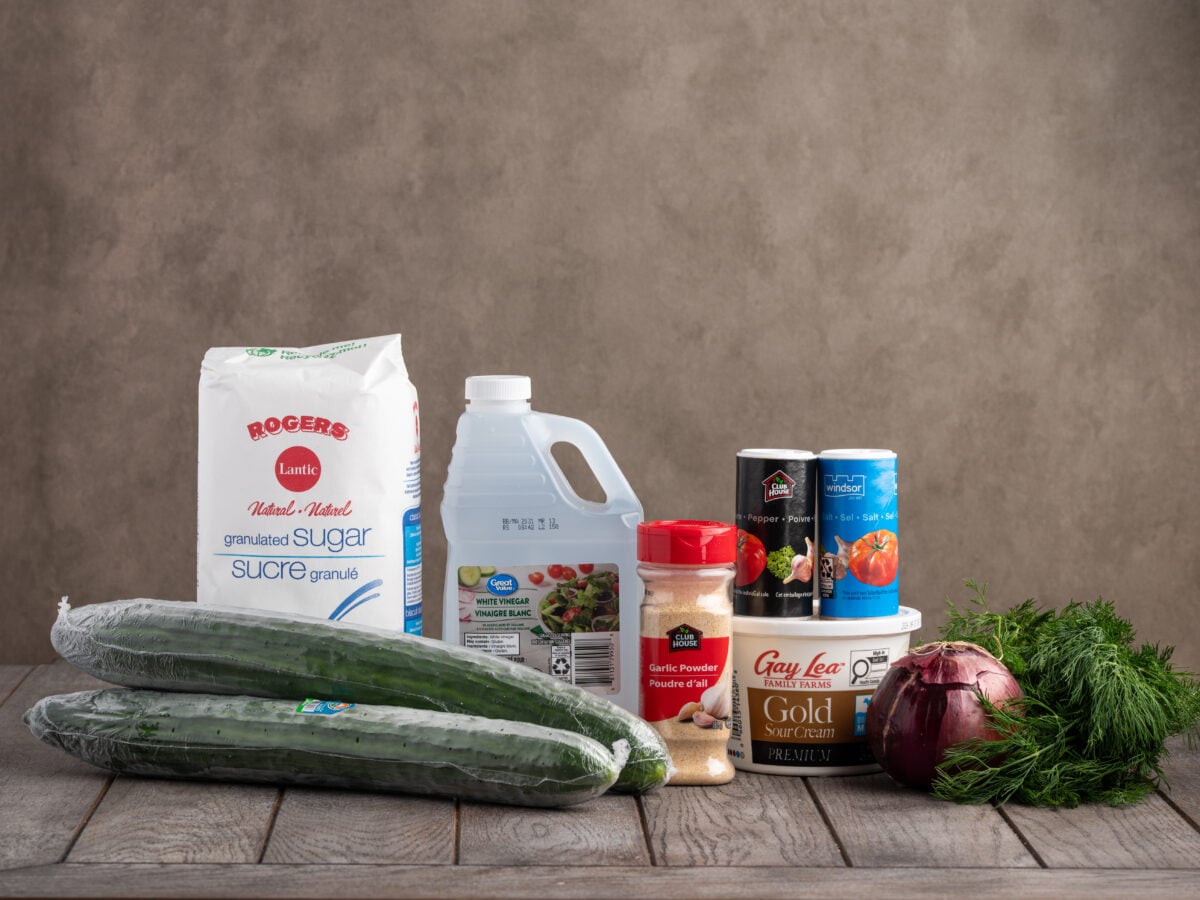 A variety of ingredients for Creamy Cucumber Salad are displayed on a wooden surface, including two cucumbers, sour cream, fresh dill, red onion, canned tomatoes, white vinegar, sugar, garlic powder, and black pepper.