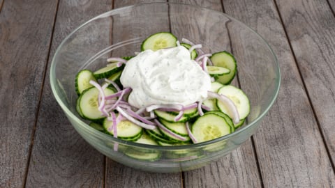 A glass bowl filled with creamy cucumber salad&mdash;sliced cucumbers and thinly sliced red onions topped with a dollop of white dressing&mdash;sits on a wooden surface.
