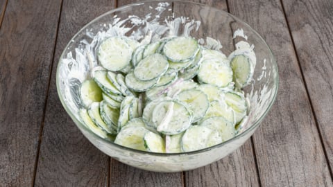 A glass bowl filled with Creamy Cucumber Salad, featuring sliced cucumbers and fresh herbs, sits on a wooden surface.