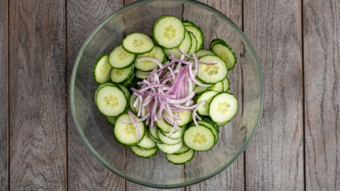 A glass bowl filled with sliced cucumbers and thinly sliced red onions, set on a rustic wooden surface&mdash;perfect for serving a refreshing Creamy Cucumber Salad.
