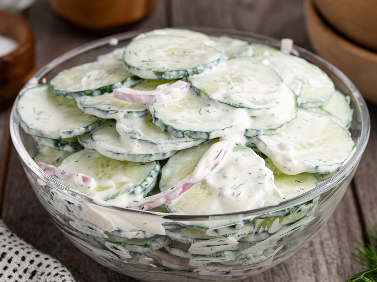 A glass bowl filled with Creamy Cucumber Salad, featuring sliced cucumbers, red onions, and a white dill dressing, sits invitingly on a wooden surface.