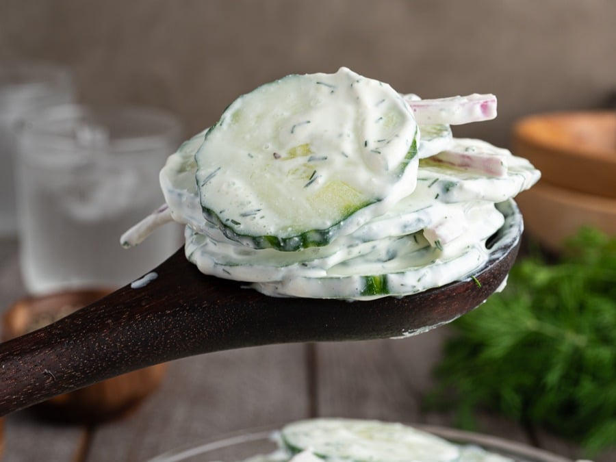 A wooden spoon holds slices of cucumber and red onion coated in a creamy, herb-filled dressing, capturing the essence of Creamy Cucumber Salad with a blurred background of fresh dill and glassware.