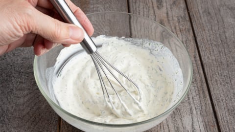 A hand using a metal whisk to mix a creamy cucumber salad dressing with herbs in a glass bowl, placed on a wooden surface.