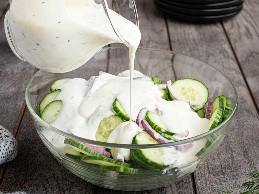 A glass bowl filled with sliced cucumbers and red onions is being topped with creamy white dressing poured from a jug, creating a classic Creamy Cucumber Salad on a wooden table.