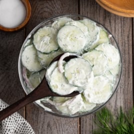 A bowl of Creamy Cucumber Salad with thinly sliced cucumbers and onions, garnished with herbs. A wooden spoon serves a portion from the rustic bowl set on a wooden table.