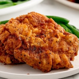 Close-up of crispy fried pork chops with a golden, craggy crust on a white plate, served with green beans in the background.