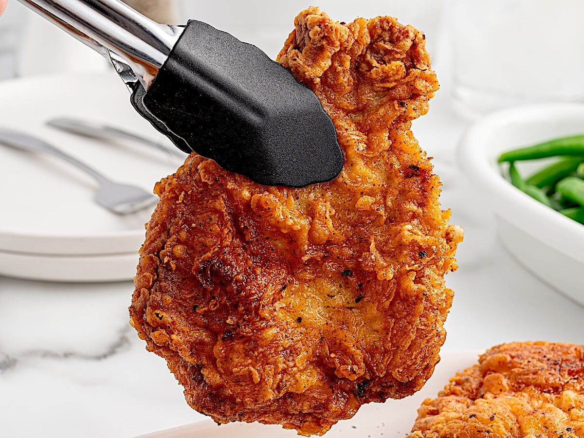 A piece of crispy fried chicken is being held by black tongs above a plate, with a fork, knife, and a bowl of green beans in the blurred background&mdash;perfect inspiration for your next Fried Pork Chops recipe.