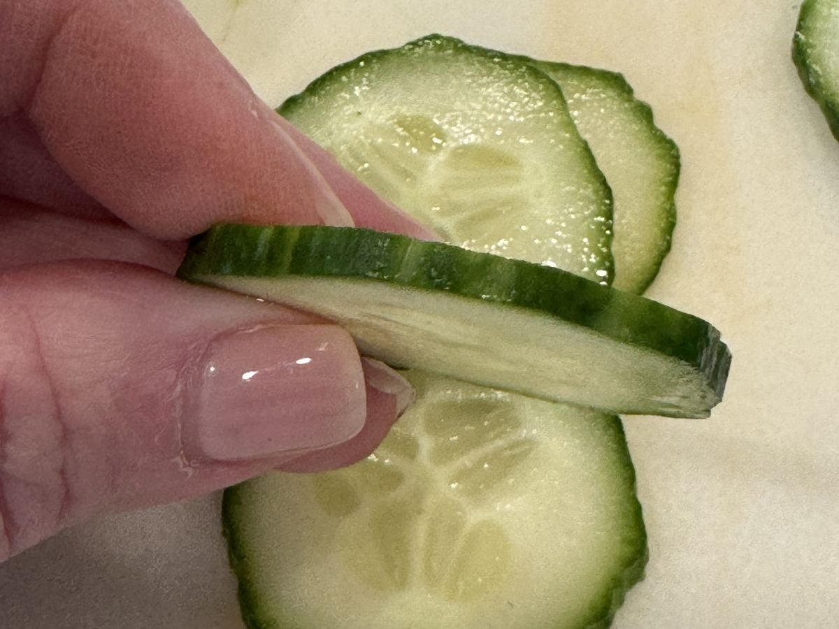 A hand with a light pink manicure holds a thin, round slice of cucumber above other cucumber slices on a light surface&mdash;perfect for preparing a fresh Creamy Cucumber Salad.