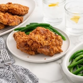 A plate with two pieces of fried chicken and green beans, next to a bowl of green beans, a platter of more fried chicken, and glasses of water with lemonโperfect inspiration for your next Fried Pork Chops Recipe on a marble table set with dotted napkins and forks.