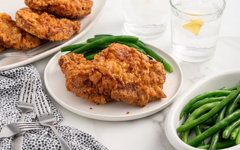 A plate with two pieces of fried chicken and green beans, next to a bowl of green beans, a platter of more fried chicken, and glasses of water with lemon—perfect inspiration for your next Fried Pork Chops Recipe on a marble table set with dotted napkins and forks.