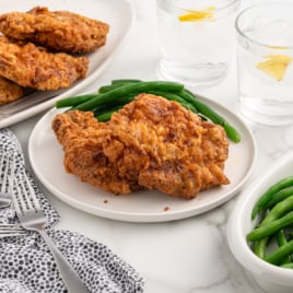 A plate with two pieces of fried chicken and green beans, plus two glasses of water with lemon. Extra fried chicken, green beans, and even some golden Fried Pork Chops appear in the background. Forks and a patterned napkin are on the side.