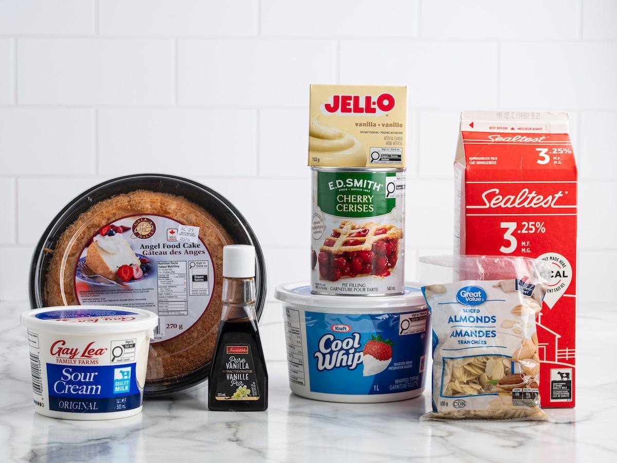A variety of dessert ingredients for a Heaven on Earth Cake sit on a marble counter: angel food cake, vanilla extract, pudding mix, cherry pie filling, milk carton, sour cream tub, whipped topping, and sliced almonds.