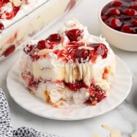 A single serving of cherry almond trifle, reminiscent of a Heaven on Earth Cake, sits on a white scalloped plate. The layered dessert features sponge cake, cream, and cherry filling, topped with maraschino cherries and sliced almonds. A bowl of cherries and extra almonds are nearby.