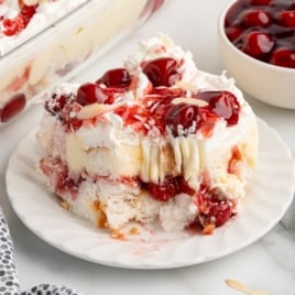 A slice of cherry-topped Heaven on Earth Cake sits with whipped cream and custard on a white plate. In the background, a pan of this irresistible Heaven on Earth Cake Recipe and a bowl of cherry topping complete the scene.