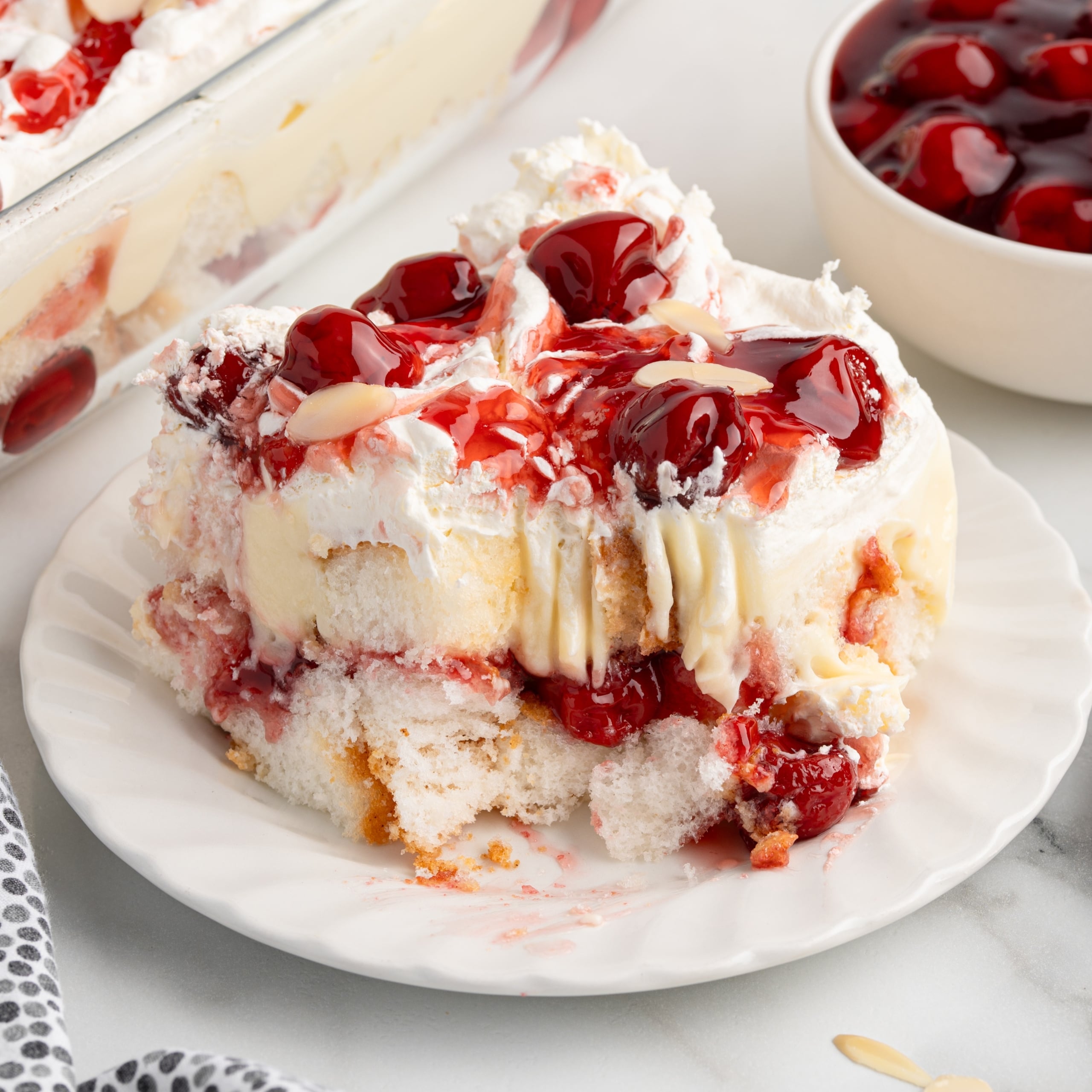 A slice of cherry-topped Heaven on Earth Cake sits with whipped cream and custard on a white plate. In the background, a pan of this irresistible Heaven on Earth Cake Recipe and a bowl of cherry topping complete the scene.