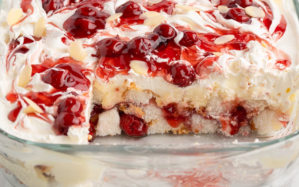 A glass baking dish filled with Heaven on Earth Cake, layered with whipped cream, cherry topping, sliced almonds, and cake layers&mdash;one corner missing to reveal the irresistible dessert&rsquo;s layers.