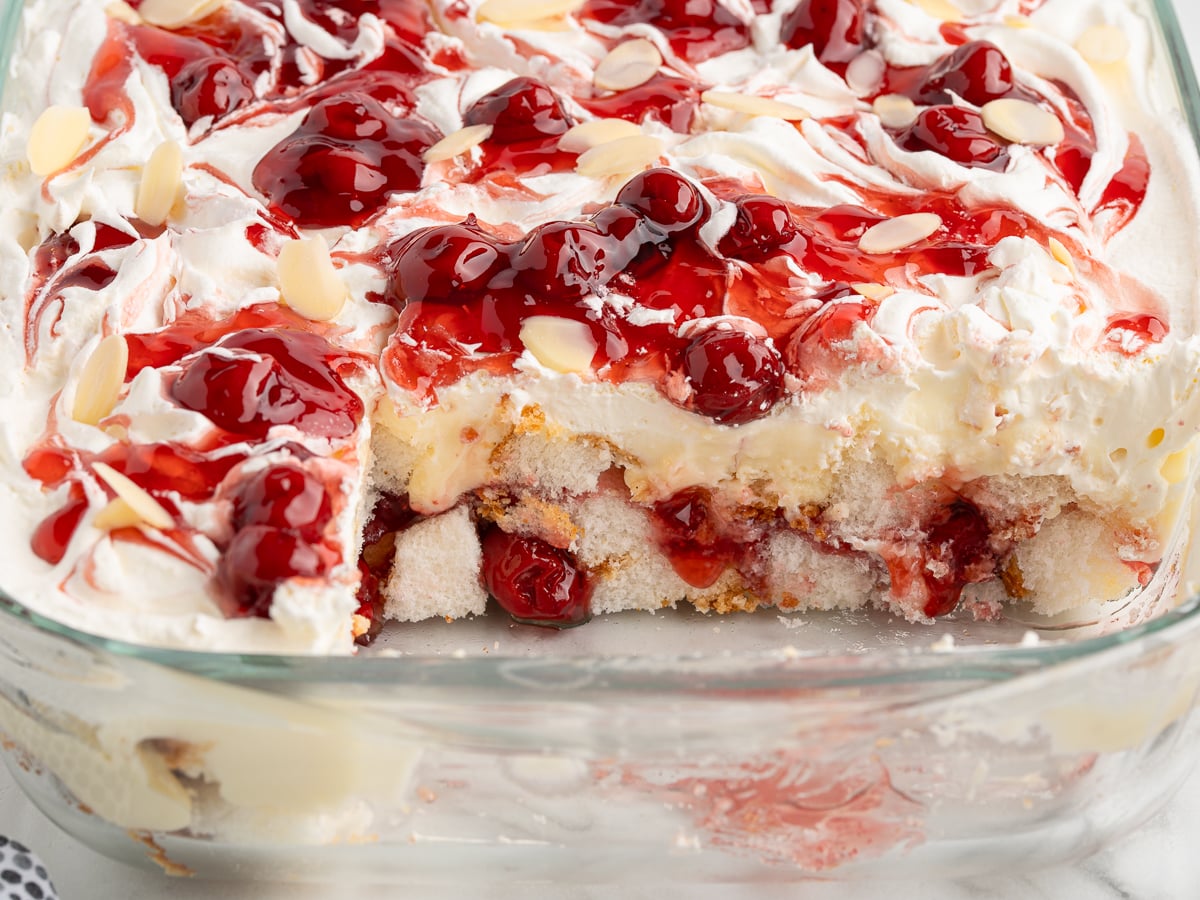 A glass baking dish filled with Heaven on Earth Cake, layered with whipped cream, cherry topping, sliced almonds, and cake layers—one corner missing to reveal the irresistible dessert’s layers.