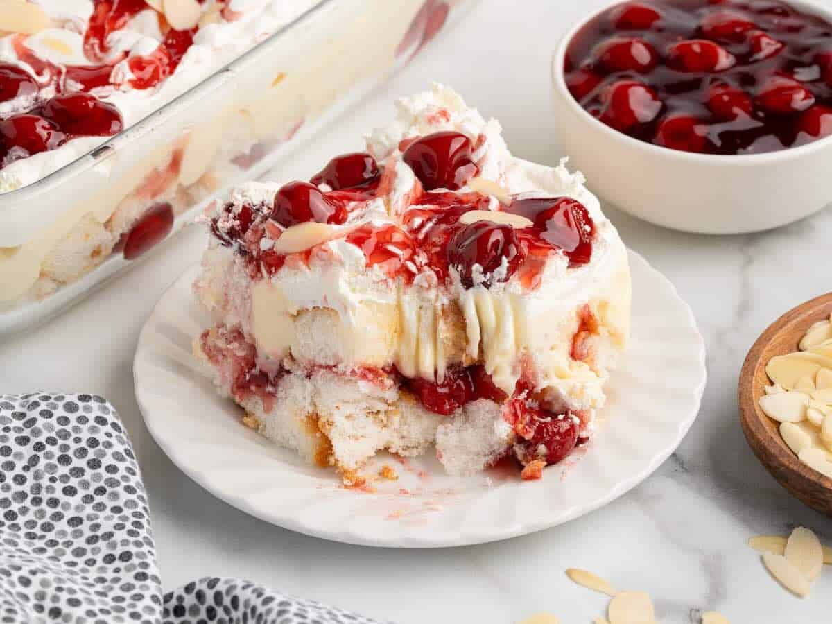 A single serving of cherry almond trifle, reminiscent of a Heaven on Earth Cake, sits on a white scalloped plate. The layered dessert features sponge cake, cream, and cherry filling, topped with maraschino cherries and sliced almonds. A bowl of cherries and extra almonds are nearby.