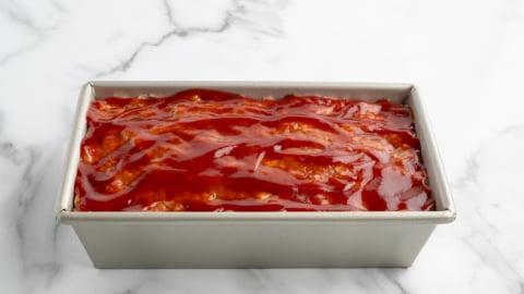 A rectangular metal loaf pan filled with uncooked meatloaf topped with a layer of ketchup sits on a white marble surface, ready to become a classic Meatloaf Recipe.