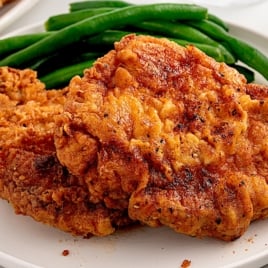 Two pieces of golden, crispy fried chicken are served on a white plate with green beans in the background, inspiring thoughts of classic comfort meals like a homemade Fried Pork Chops Recipe.