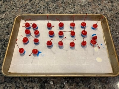 A baking sheet lined with parchment paper holds 20 4th of July Cherry Bombs, each maraschino cherry dipped in blue coating at the base, spaced evenly across the sheet on a granite countertop.