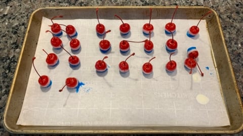 A baking sheet lined with parchment paper holds 20 4th of July Cherry Bombs, each maraschino cherry dipped in blue coating at the base, spaced evenly across the sheet on a granite countertop.