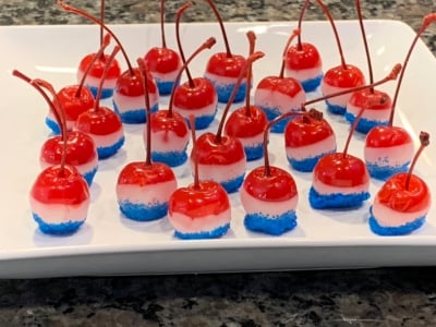 Red cherries with stems are dipped in white and blue layers, arranged on a white rectangular plate as festive 4th of July Cherry Bombs. The blue coating looks like sugar or sprinkles, making them perfect patriotic treats.
