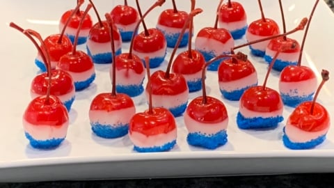 Red cherries with stems are dipped in white and blue layers, arranged on a white rectangular plate as festive 4th of July Cherry Bombs. The blue coating looks like sugar or sprinkles, making them perfect patriotic treats.
