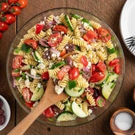 A bowl of Greek Pasta Salad made with rotini, tomatoes, cucumbers, olives, feta cheese, and fresh herbs sits on a wooden table. Nearby are plates, forks, seasonings, a tomato vine, and small bowls of olives and crumbled feta—everything you need for a refreshing Greek Salad Recipe.
