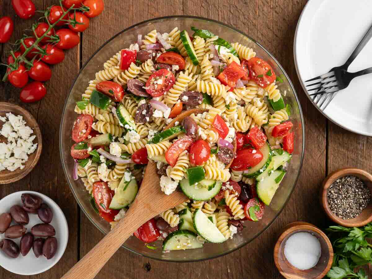 A bowl of Greek Pasta Salad made with rotini, tomatoes, cucumbers, olives, feta cheese, and fresh herbs sits on a wooden table. Nearby are plates, forks, seasonings, a tomato vine, and small bowls of olives and crumbled feta—everything you need for a refreshing Greek Salad Recipe.