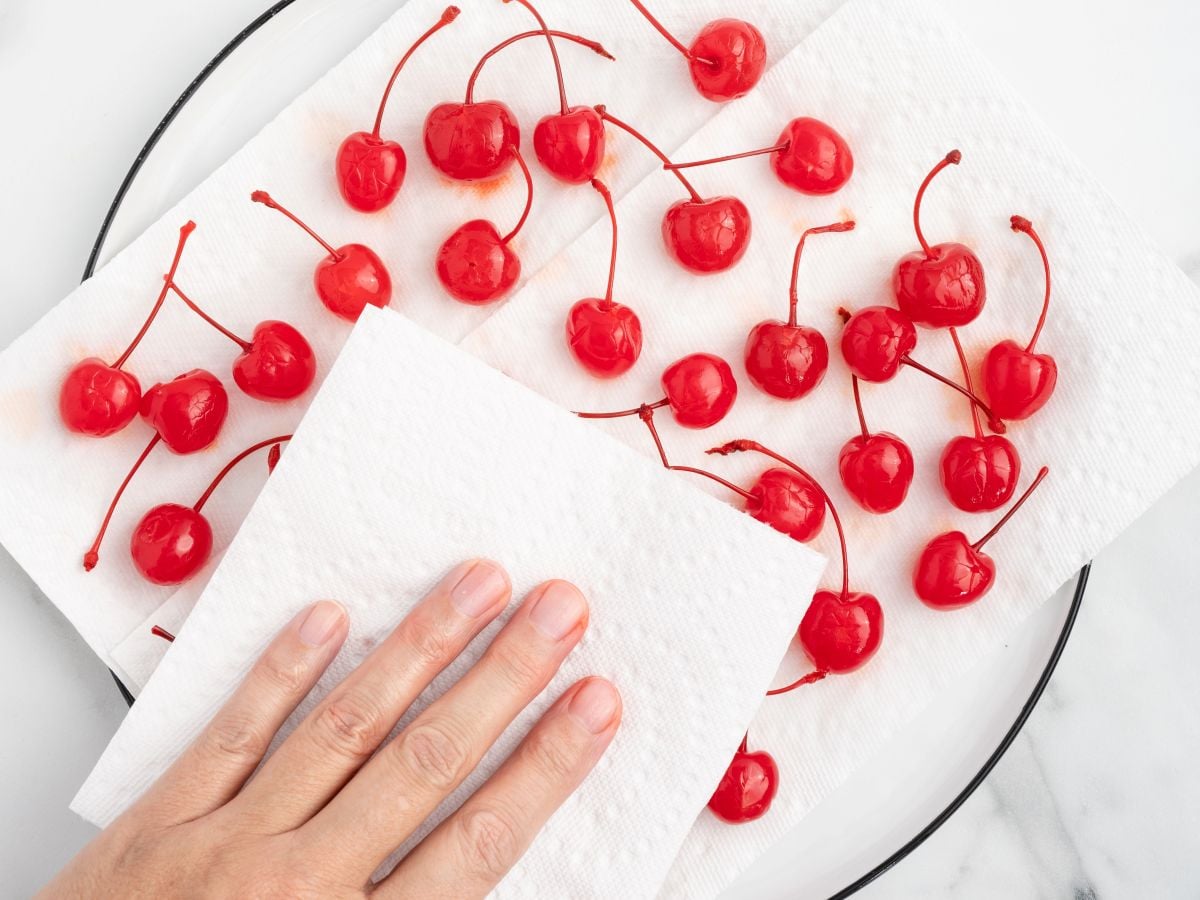 A hand presses a paper towel over several bright red maraschino cherries with stems, arranged on another paper towel on a plate&mdash;perfect prep for making 4th of July Cherry Bombs.