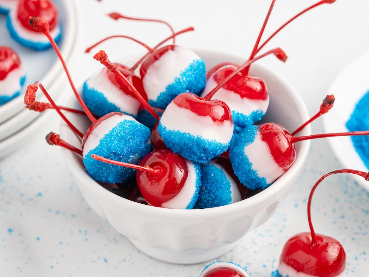 A white bowl filled with 4th of July Cherry Bombs&mdash;cherries coated in white and blue sugar layers, with long stems, perfectly decorated in a festive red, white, and blue color scheme.