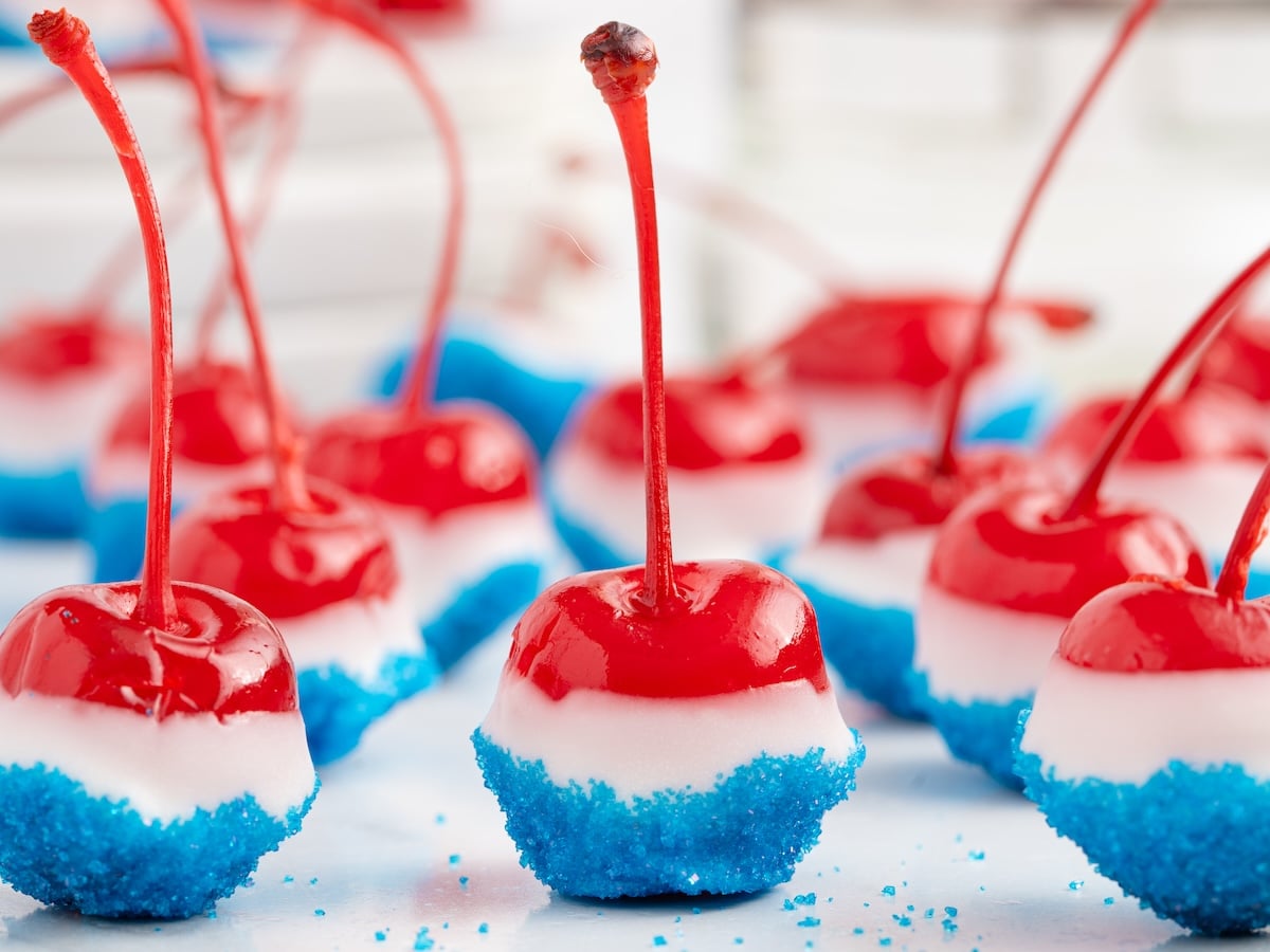 Several 4th of July Cherry Bombs&mdash;maraschino cherries dipped in white candy coating and blue sugar&mdash;are arranged on a white surface, creating a festive red, white, and blue patriotic treat.