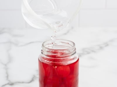 A clear liquid is being poured from a glass measuring cup into a jar filled with red liquid and whole cherries—perfect for making festive 4th of July Cherry Bombs on a white marble surface.