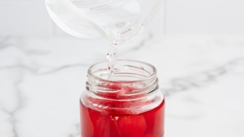 A clear liquid is being poured from a glass measuring cup into a jar filled with red liquid and whole cherries&mdash;perfect for making festive 4th of July Cherry Bombs on a white marble surface.