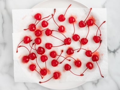 A cluster of bright red 4th of July Cherry Bombs with stems rests on paper towels atop a white plate, on a marble surface. Some cherry juice has stained the paper towels.