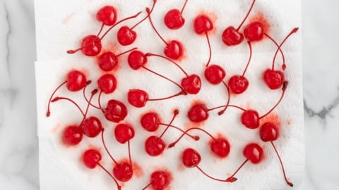A cluster of bright red 4th of July Cherry Bombs with stems rests on paper towels atop a white plate, on a marble surface. Some cherry juice has stained the paper towels.