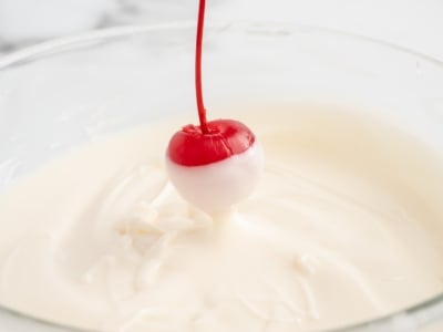A maraschino cherry on a stem is being dipped halfway into a bowl of creamy white chocolate, creating the perfect look for festive 4th of July Cherry Bombs. The coating is clearly seen on the lower half of the cherry.
