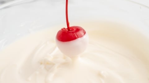 A maraschino cherry on a stem is being dipped halfway into a bowl of creamy white chocolate, creating the perfect look for festive 4th of July Cherry Bombs. The coating is clearly seen on the lower half of the cherry.