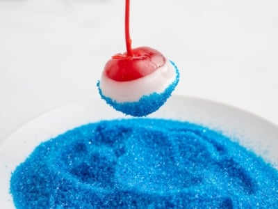 A maraschino cherry dipped in white chocolate is being rolled in bright blue sugar crystals, creating festive 4th of July Cherry Bombs that hover above a plate filled with the same sparkling blue sugar.
