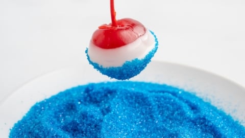 A maraschino cherry dipped in white chocolate is being rolled in bright blue sugar crystals, creating festive 4th of July Cherry Bombs that hover above a plate filled with the same sparkling blue sugar.