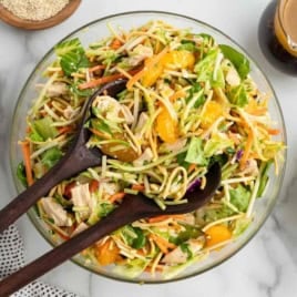 A glass bowl of Asian-inspired Chinese Chicken Salad with lettuce, shredded carrots, broccoli slaw, mandarin oranges, and chunks of chicken, served with two wooden salad servers; dressing and green onions are on the side.