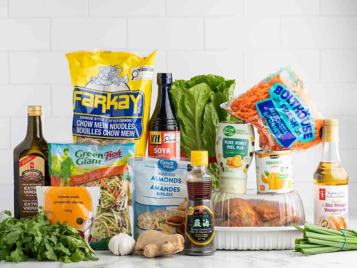 A variety of groceries for a delicious Chinese Chicken Salad, including chow mein noodles, soy sauce, vegetables, chicken, oils, rice vinegar, almonds, and sauces are arranged on a counter against a white tile background.