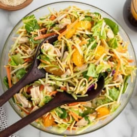 A bowl of Asian-inspired salad with greens, shredded carrots, mandarin oranges, chicken, and crunchy noodles invites you to enjoy a fresh take on a classic Chinese Chicken Salad Recipe. Wooden salad servers rest on top, ready for tossing and serving this delicious Chinese Chicken Salad.