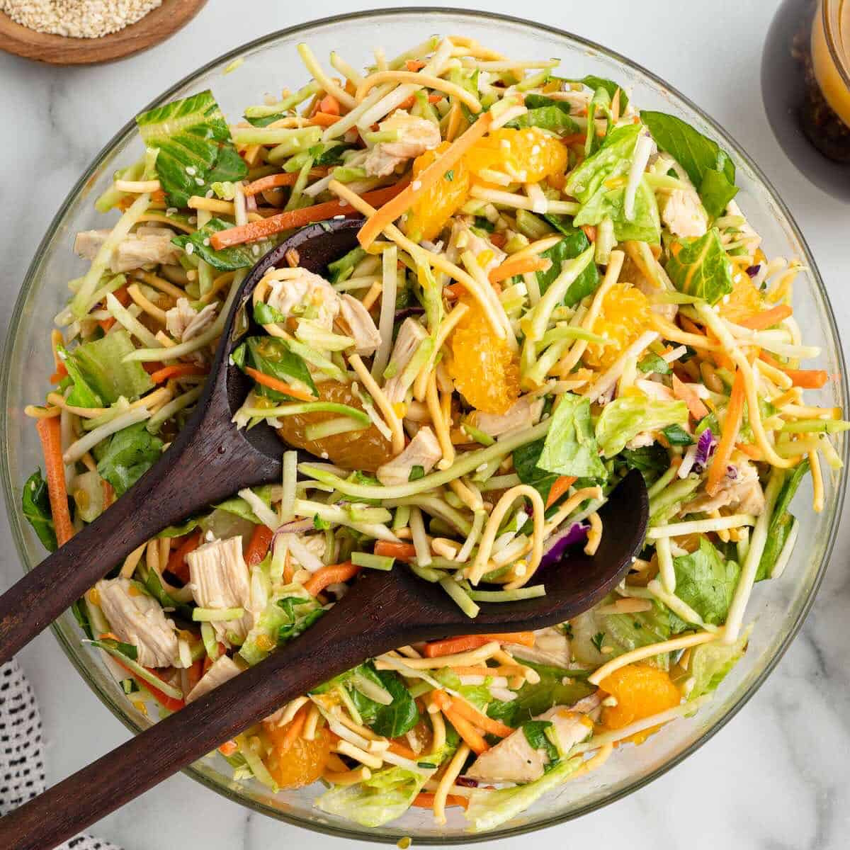 A bowl of Asian-inspired salad with greens, shredded carrots, mandarin oranges, chicken, and crunchy noodles invites you to enjoy a fresh take on a classic Chinese Chicken Salad Recipe. Wooden salad servers rest on top, ready for tossing and serving this delicious Chinese Chicken Salad.
